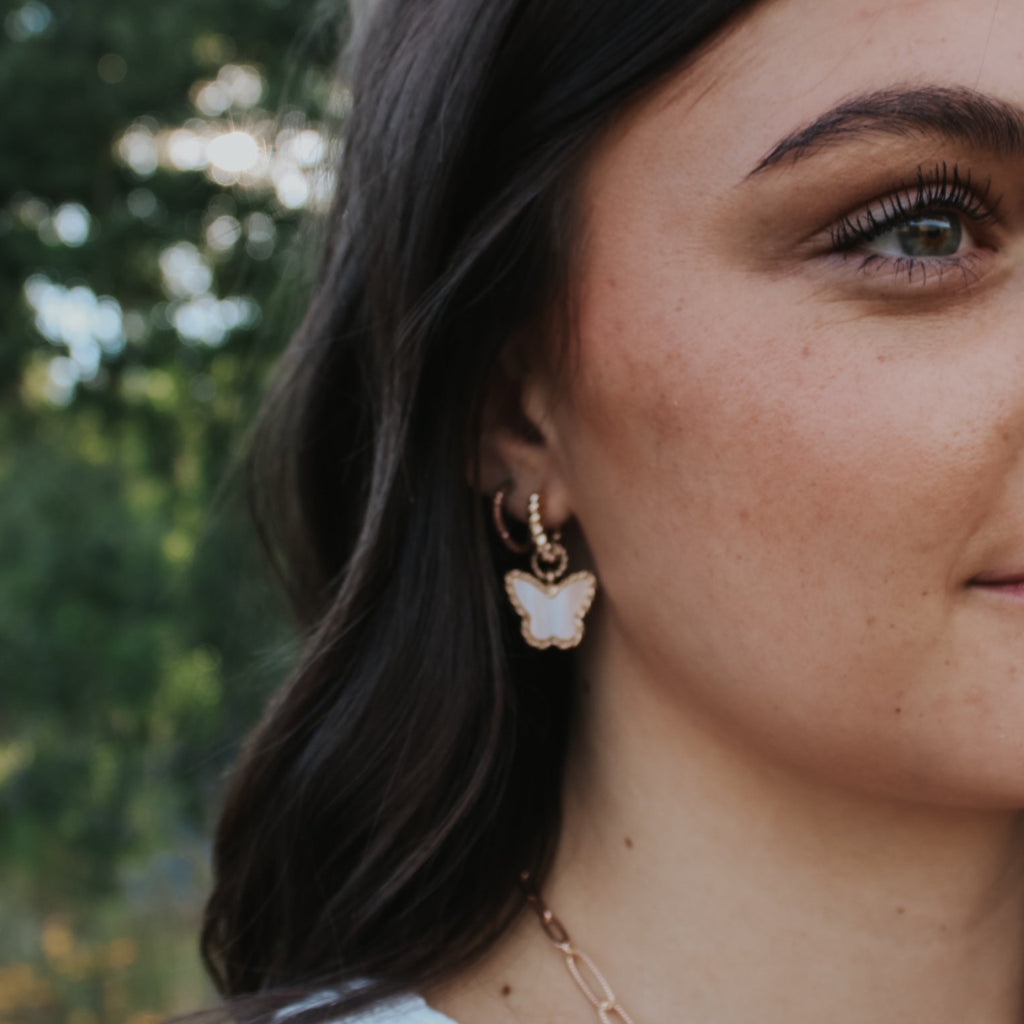 Woman with dark hair and earrings standing in a natural setting with trees and plants.