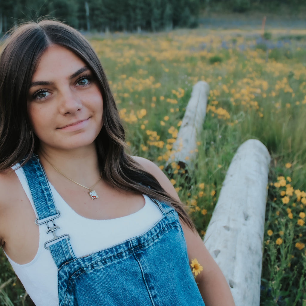 Woman in a field of yellow flowers wearing denim overalls. pendant necklace. cherries gold